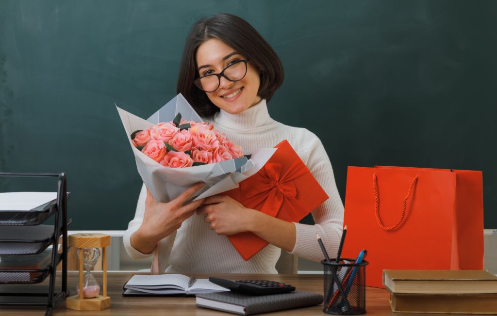 smiling-young-female-teacher-holding-flowers-with-gift-sitting-desk-with-school-tools-classroom2.jpg